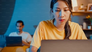 Freelance asia couple man and woman in casual wife focus financial work business in laptop computer on desk at night happy husband video call with family rest lay down after work on couch at home.