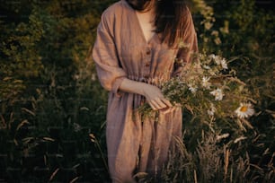 화창한 저녁에 여름 초원에서 야생화를 모으는 린넨 드레스를 입은 아름다운 여자가 문을 닫습니다. 대기 순간. 시골에서 꽃을 따는 소박한 드레스를 입은 세련된 젊은 여성. 느린 삶