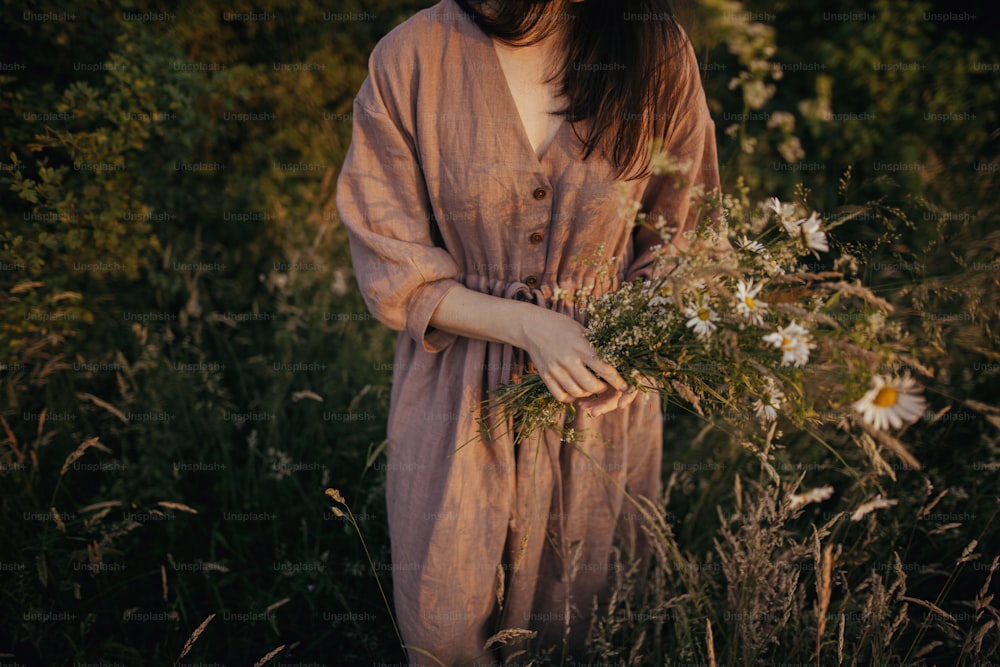 Schöne Frau in Leinenkleid sammelt Wildblumen auf Sommerwiese am sonnigen Abend, Nahaufnahme. Atmosphärischer Moment. Stilvolle junge Frau in rustikalem Kleid, die Blumen auf dem Land pflückt. Langsames Leben