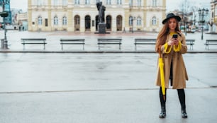 Junge schöne Frau, die ein Smartphone benutzt und einen gelben Regenschirm hält, während sie an einem regnerischen Tag draußen in der Stadt ist