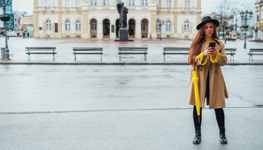 Young beautiful woman using a smartphone and holding a yellow umbrella while outside in the city on a rainy day