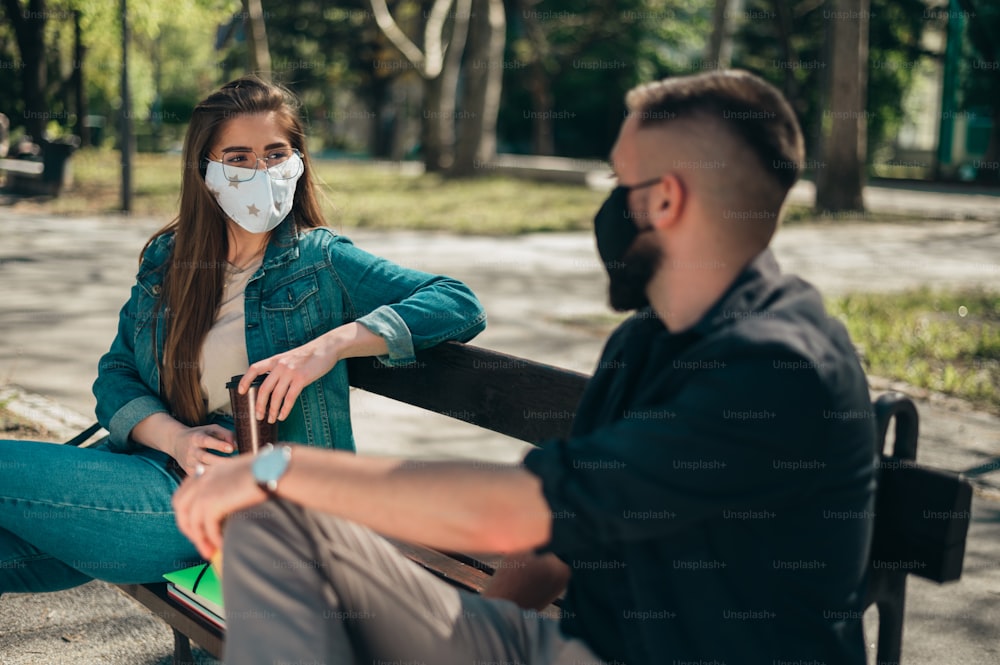 Due giovani studenti che indossano maschere protettive e si siedono su una panchina in un campus e prendono le distanze a causa di una pandemia di coronavirus