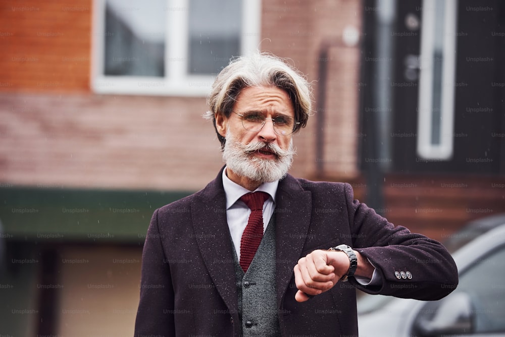 Fashionable senior man with gray hair and beard is outdoors on the street near his car cheks time on his watch.