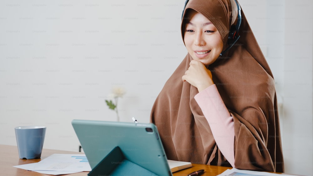 아시아 무슬림 여성은 디지털 태블릿을 사용하여 헤드폰을 착용하고 부엌에서 집에서 일하는 동안 회의 화상 통화에서 판매 보고서에 대해 동료들과 이야기합니다. 사회적 거리두기, 코로나 바이러스 격리.
