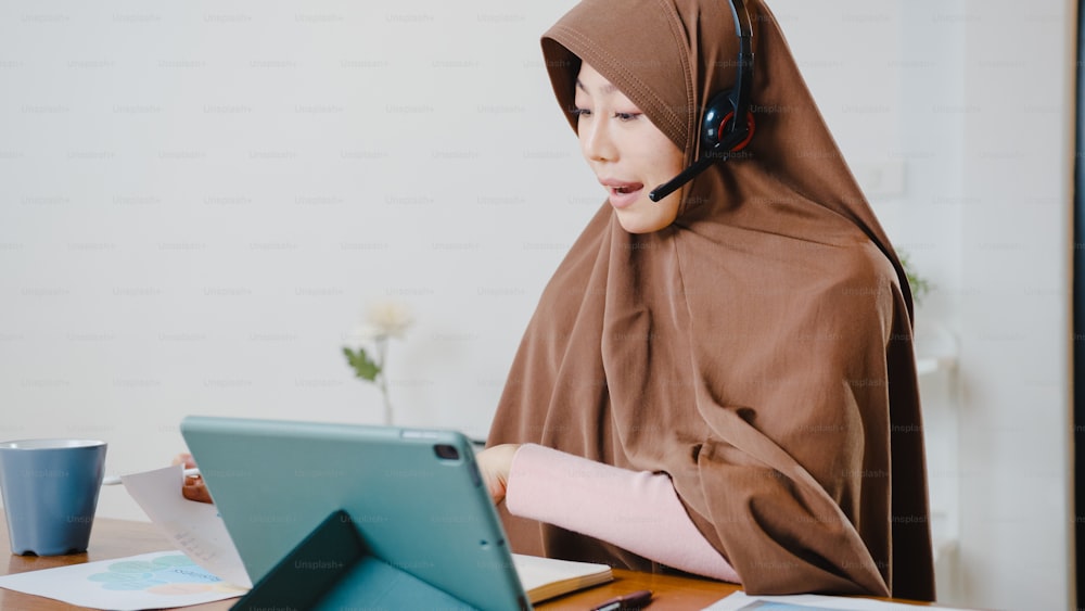 アジアのイスラム教徒の女性は、キッチンで自宅で仕事をしながら、デジタルタブレットを使用してヘッドフォンを着用し、会議のビデオ通話で販売レポートについて同僚と話します。ソーシャルディスタンス、コロナウイルスの検疫。
