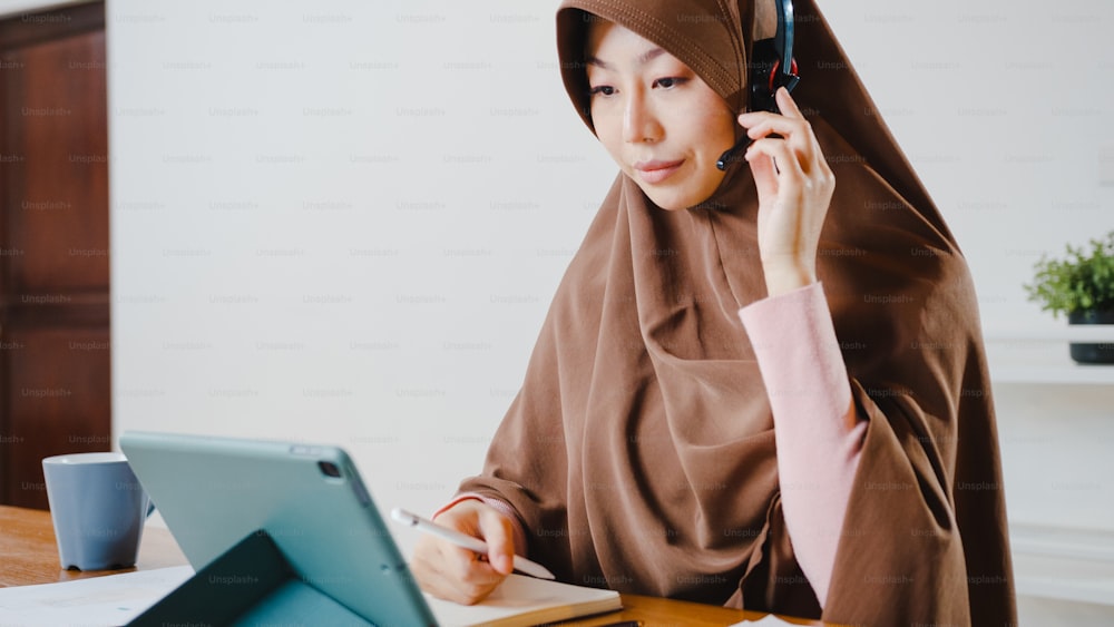 アジアのイスラム教徒の女性は、キッチンで自宅で仕事をしながら、デジタルタブレットを使用してヘッドフォンを着用し、会議のビデオ通話で販売レポートについて同僚と話します。ソーシャルディスタンス、コロナウイルスの検疫。