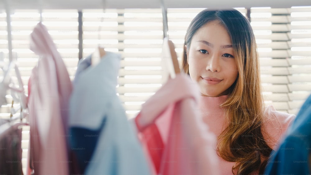 Bella giovane donna asiatica attraente che sceglie i suoi vestiti di vestito di moda nell'armadio a casa o in negozio. La ragazza pensa a cosa indossare la camicia casual. Guardaroba di casa o spogliatoio del negozio di abbigliamento.