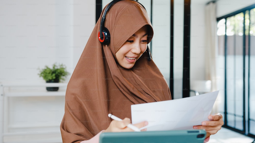 Senhora muçulmana da Ásia usa fone de ouvido usando tablet digital converse com colegas sobre relatório de venda em chamada de vídeo em conferência enquanto trabalha em casa na cozinha. Distanciamento social, quarentena para corona vírus.