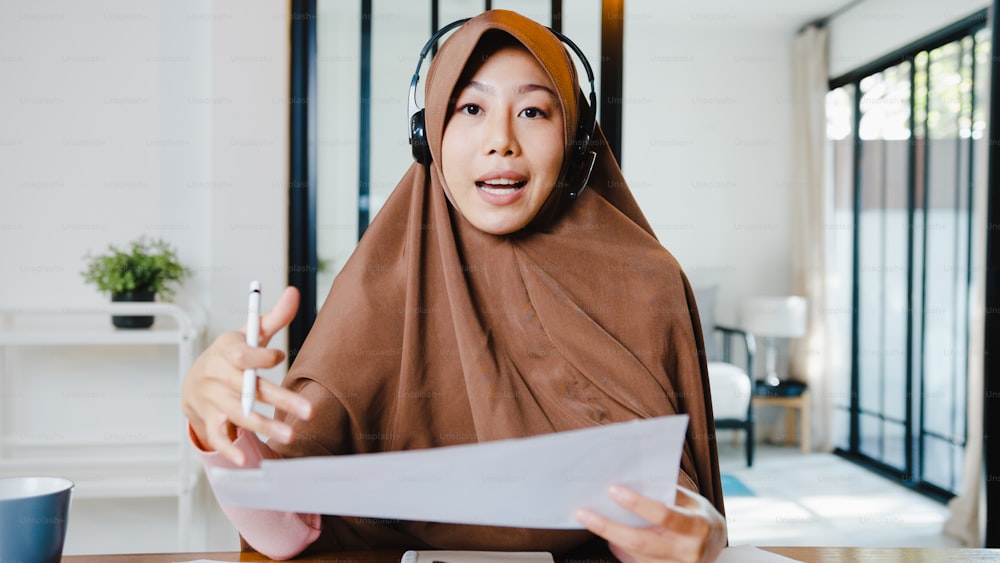 アジアのイスラム教徒の女性は、自宅のリビングルームでリモートワークをしながら、コンピューターのラップトップを使用してヘッドフォンを着用し、ビデオ通話で販売レポートについて同僚と話します。ソーシャルディスタンス、コロナウイルスの検疫。