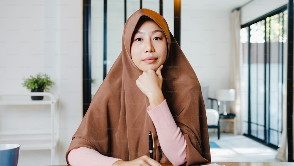 La signora musulmana asiatica indossa l'hijab usando il computer portatile parla con i colleghi del piano in videochiamata mentre lavora a distanza da casa in salotto. Distanziamento sociale, quarantena per il corona virus.