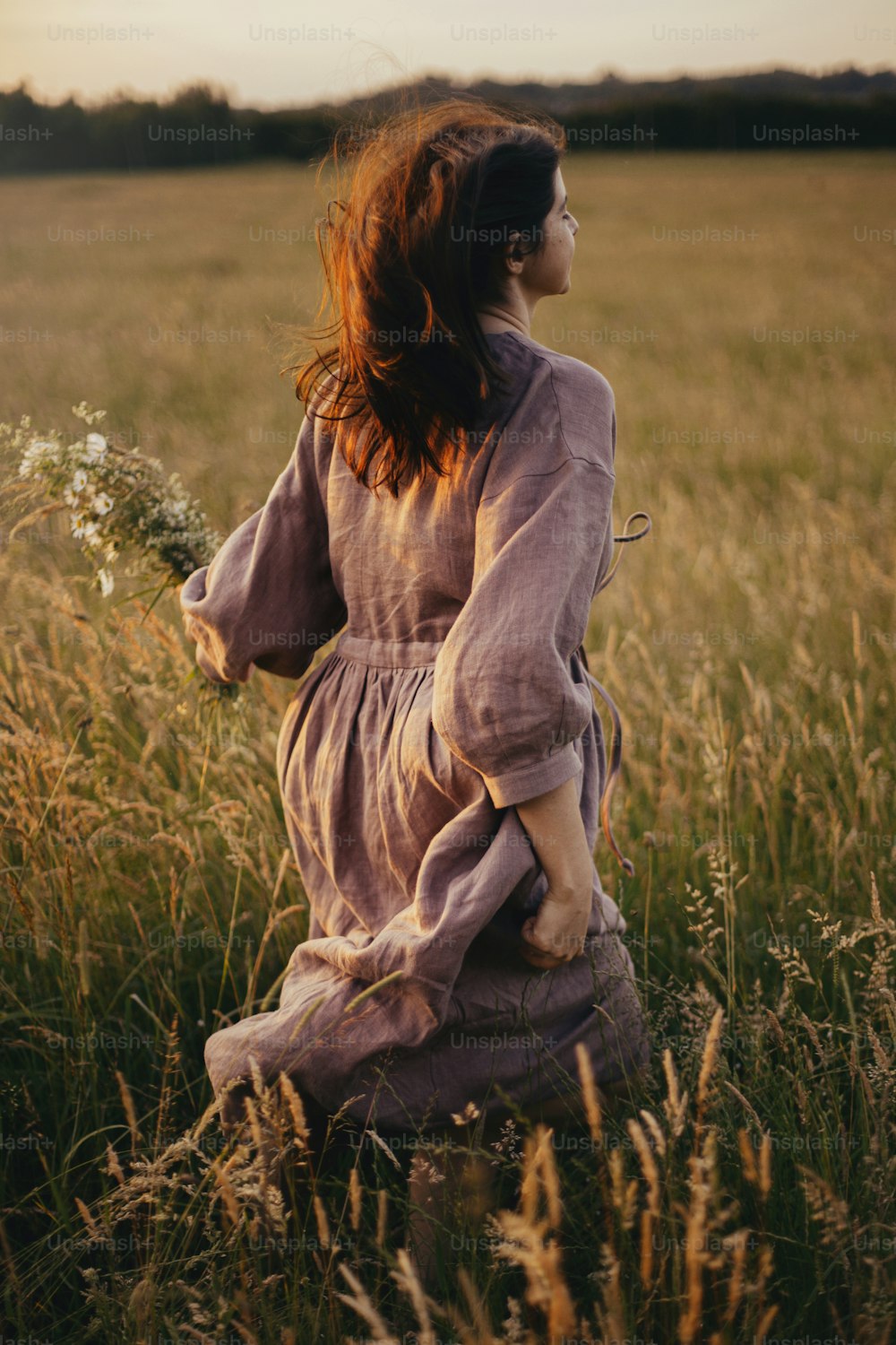 Mulher bonita no vestido de linho correndo com flores silvestres na mão no prado de verão no pôr do sol. Elegante jovem fêmea em vestido rústico desfrutando de noite livre no campo. Momento despreocupado atmosférico