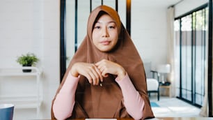 La signora musulmana asiatica indossa l'hijab usando il computer portatile parla con i colleghi del piano in videochiamata mentre lavora a distanza da casa in salotto. Distanziamento sociale, quarantena per il corona virus.