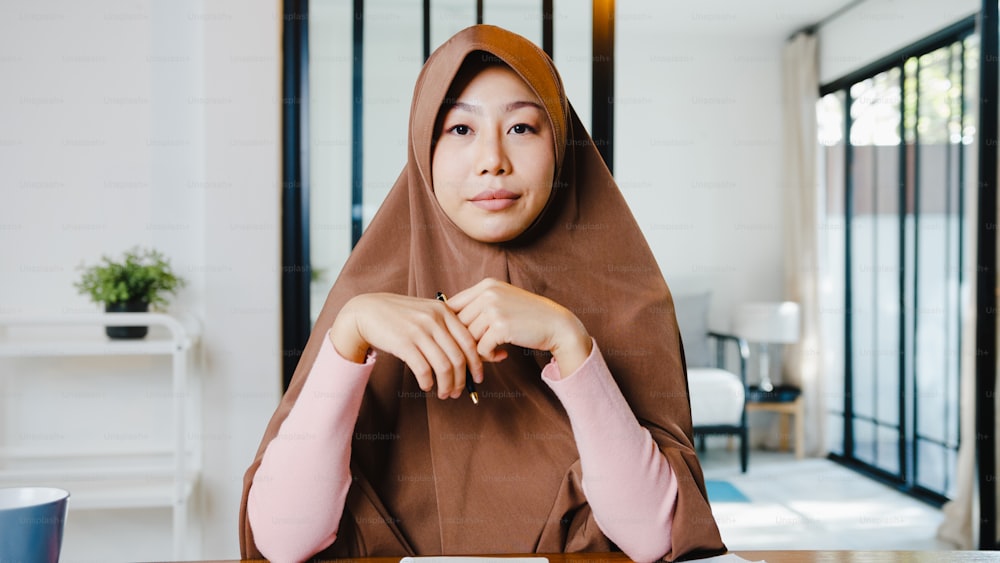 アジアのイスラム教徒の女性は、自宅のリビングルームでリモートワークをしながら、コンピューターのラップトップを使用してヒジャーブを着用し、ビデオ通話会議で計画について同僚と話します。ソーシャルディスタンス、コロナウイルスの検疫。