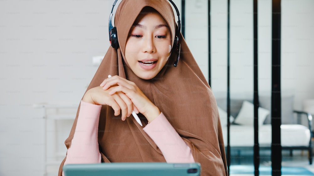 Asia muslim lady wear headphone using digital tablet talk to colleagues about sale report in conference video call while working from home at kitchen. Social distancing, quarantine for corona virus.