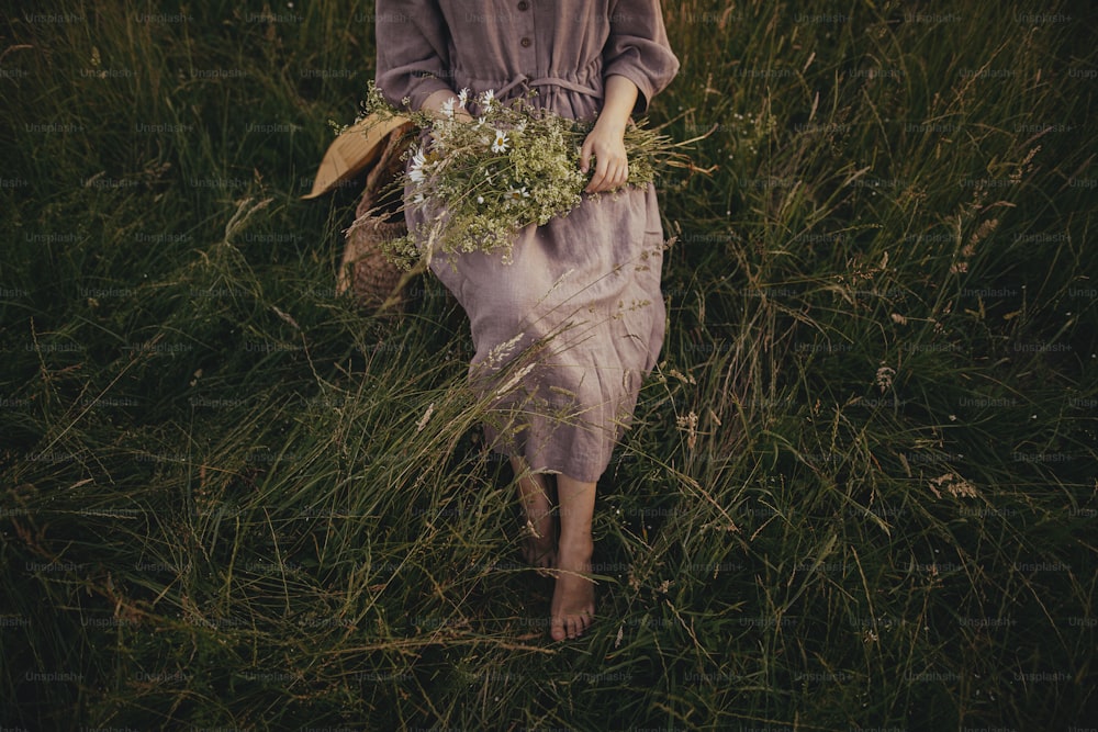 리넨 드레스를 입은 아름다운 여자가 여름 초원에서 야생화 꽃다발과 함께 잔디 사이의 소박한 의자에 맨발로 앉아 있다. 시골에서 휴식을 취하는 젊은 여성. 대기 순간. 상위 뷰