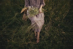 Beautiful woman in linen dress sitting barefoot on rustic chair among grass with bouquet of wildflowers in summer meadow. Young female relaxing in countryside. Atmospheric moment. Top view
