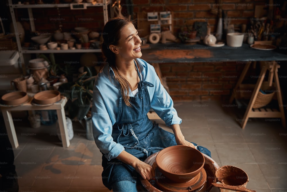 Donna artigianale positiva che indossa un grembiule seduta vicino al tornio di ceramica mentre distoglie lo sguardo durante il processo di produzione della ciotola di ceramica nello studio d'arte