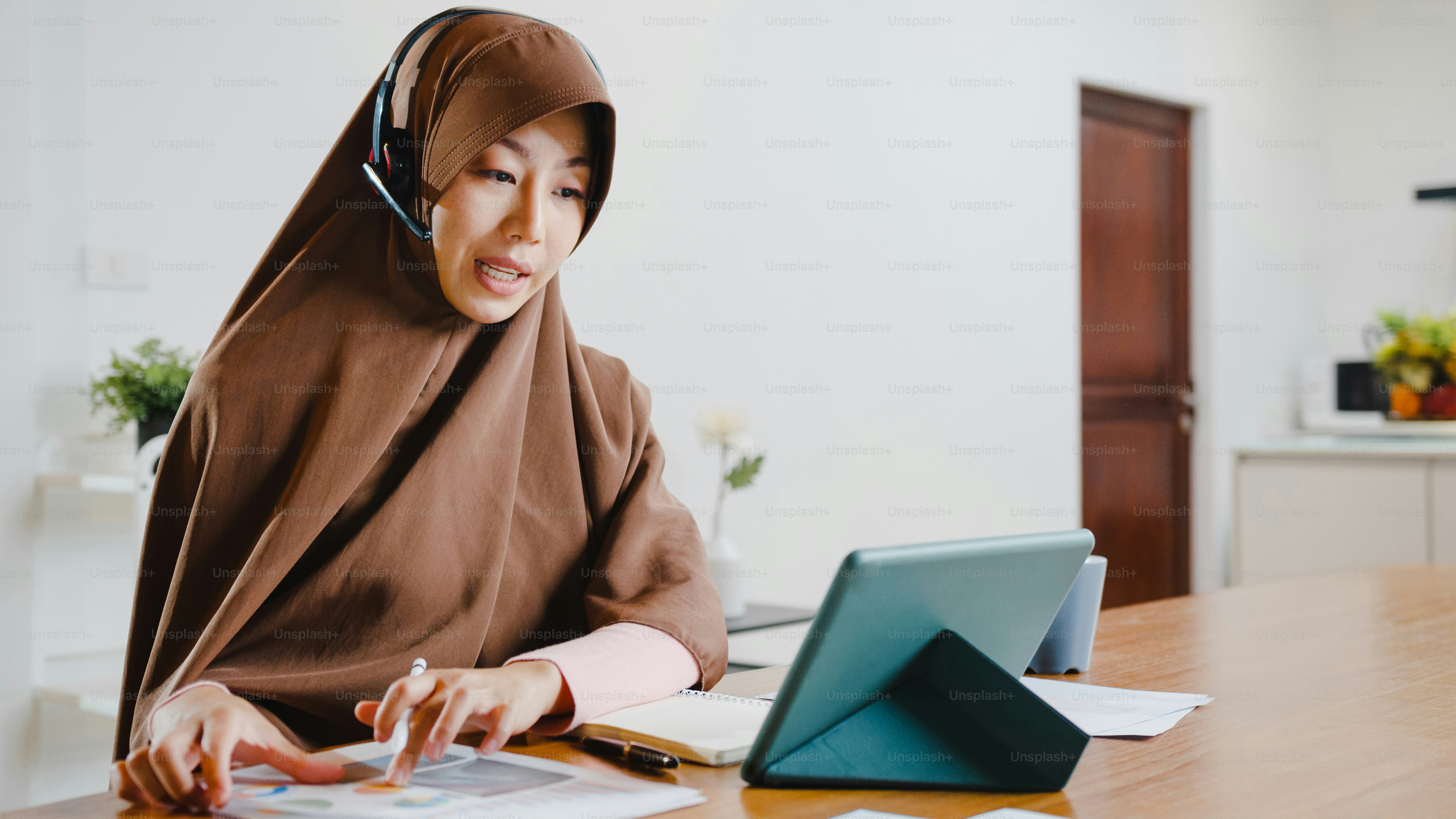 Asia muslim lady wear headphone using digital tablet talk to colleagues about sale report in conference video call while working from home at kitchen. Social distancing, quarantine for corona virus.