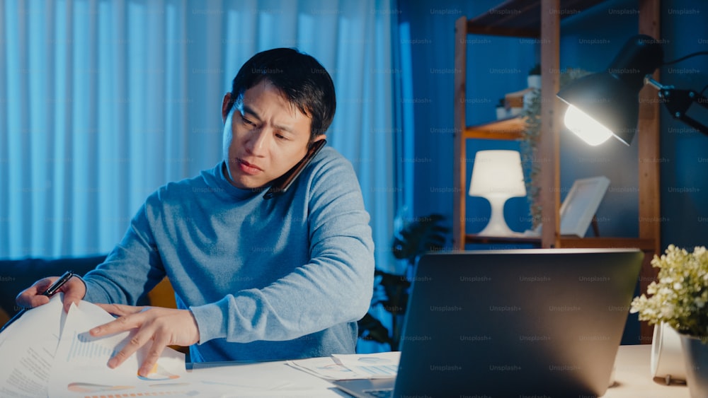 Young Asia businessman use smartphone call meeting agenda assignment paperwork with colleague look at laptop computer in living room at home overtime at night, Work from home corona pandemic concept.