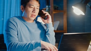 Junge asiatische Geschäftsmann verwenden Smartphone-Anruf Treffen Tagesordnung Zuweisung Papierkram mit Kollegen Blick auf Laptop-Computer im Wohnzimmer zu Hause Überstunden in der Nacht, Arbeit von zu Hause Corona-Pandemie-Konzept.