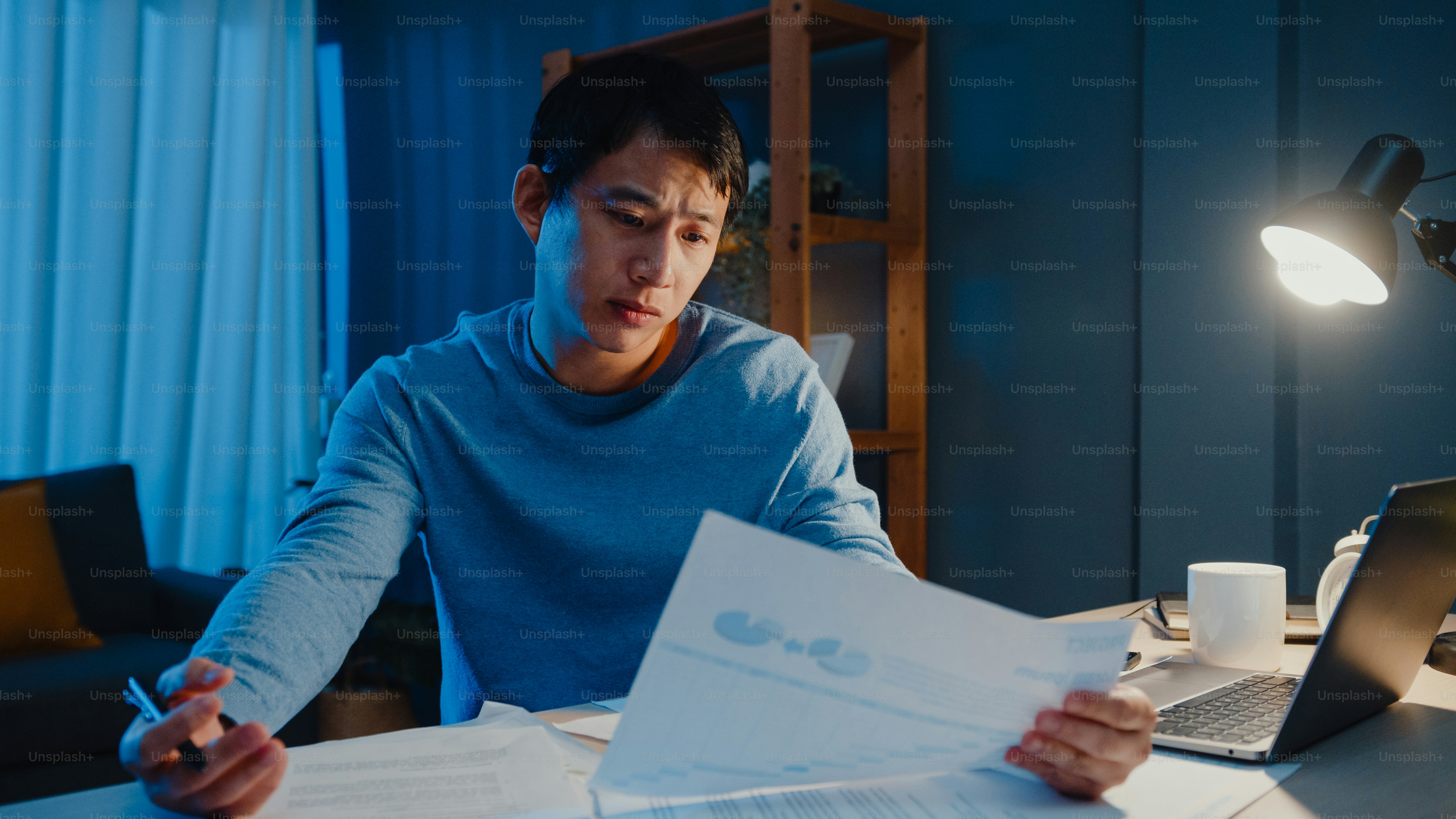 Asian freelance businessman focus work type on laptop computer busy with full of paperwork chart on desk in living room at home overtime at night, Work from home during COVID-19 pandemic concept.