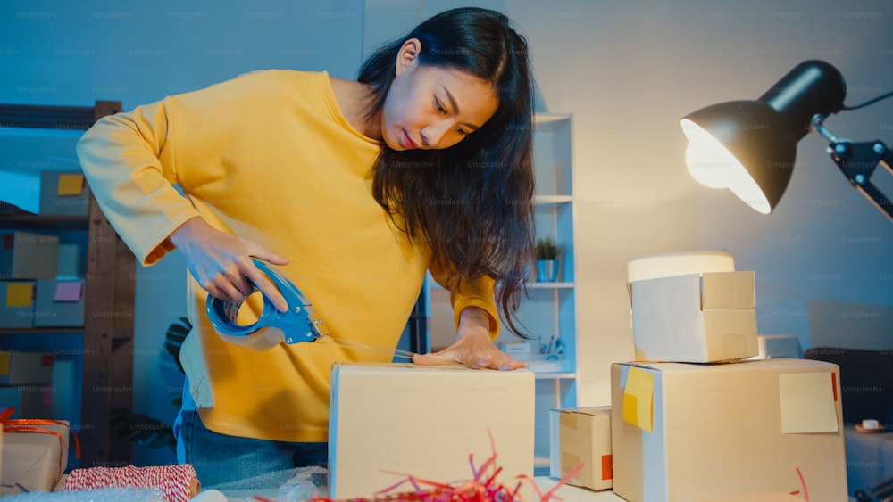 Young Asia businesswoman prepare product use tape packing box for send to customer purchase order in home office at night. Small business owner, online market delivery, lifestyle freelance concept.