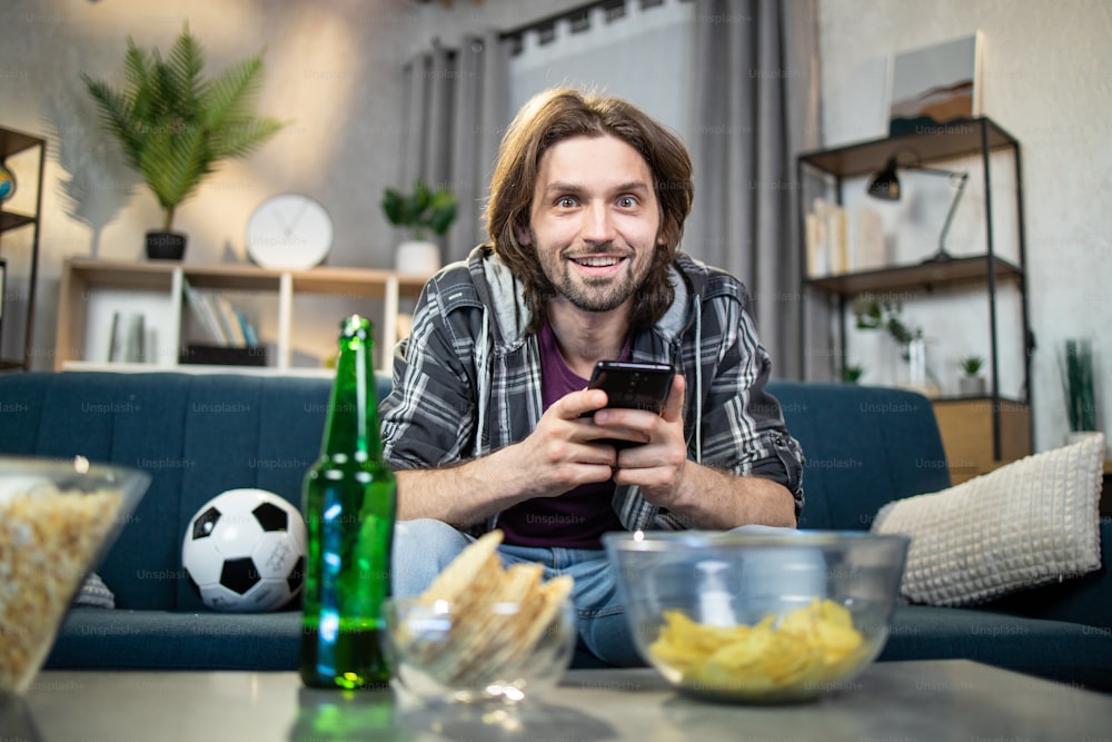 Glücklicher, gutaussehender Mann mit modernem Smartphone, um Live-Stream der Fußball-Europameisterschaft zu sehen. Aufgeregter Typ mit Bier und Snacks, der die Lieblingsfußballmannschaft unterstützt.