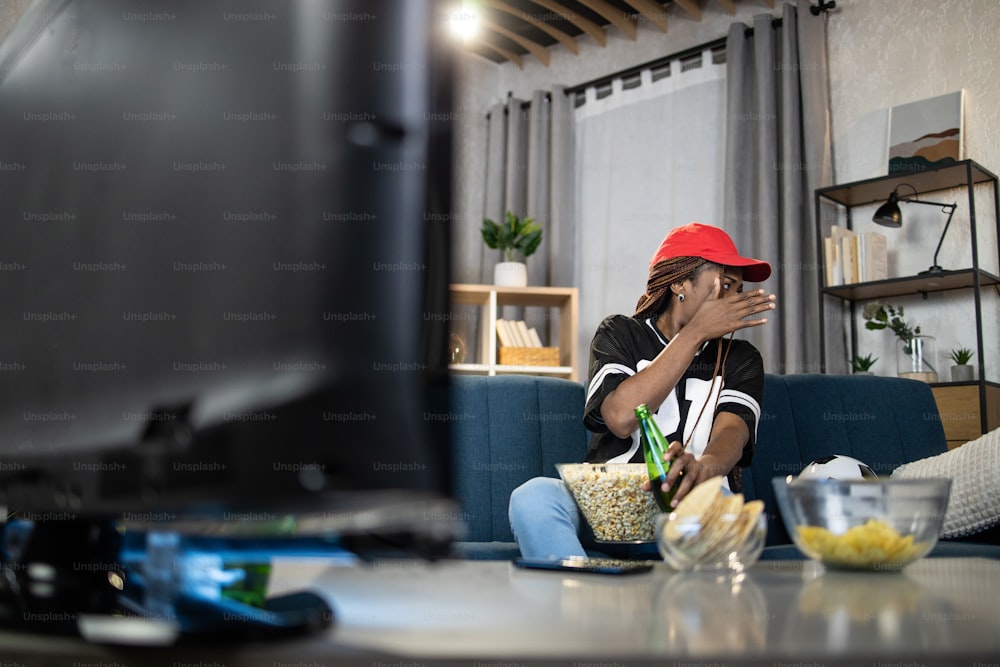 Donna attraente che guarda da parte e copre il viso con il palmo della mano durante il momento emotivo della partita di calcio. Femmina africana seduta a casa e guardando la TV.