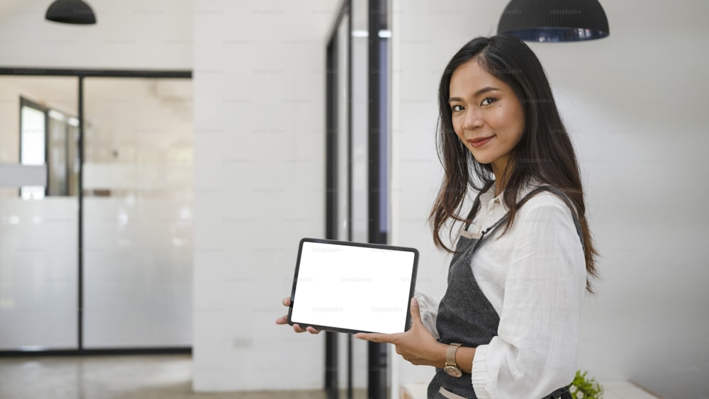 Friendly female coffee shop owner in apron showing digital tablet. Blank screen for advertising text.
