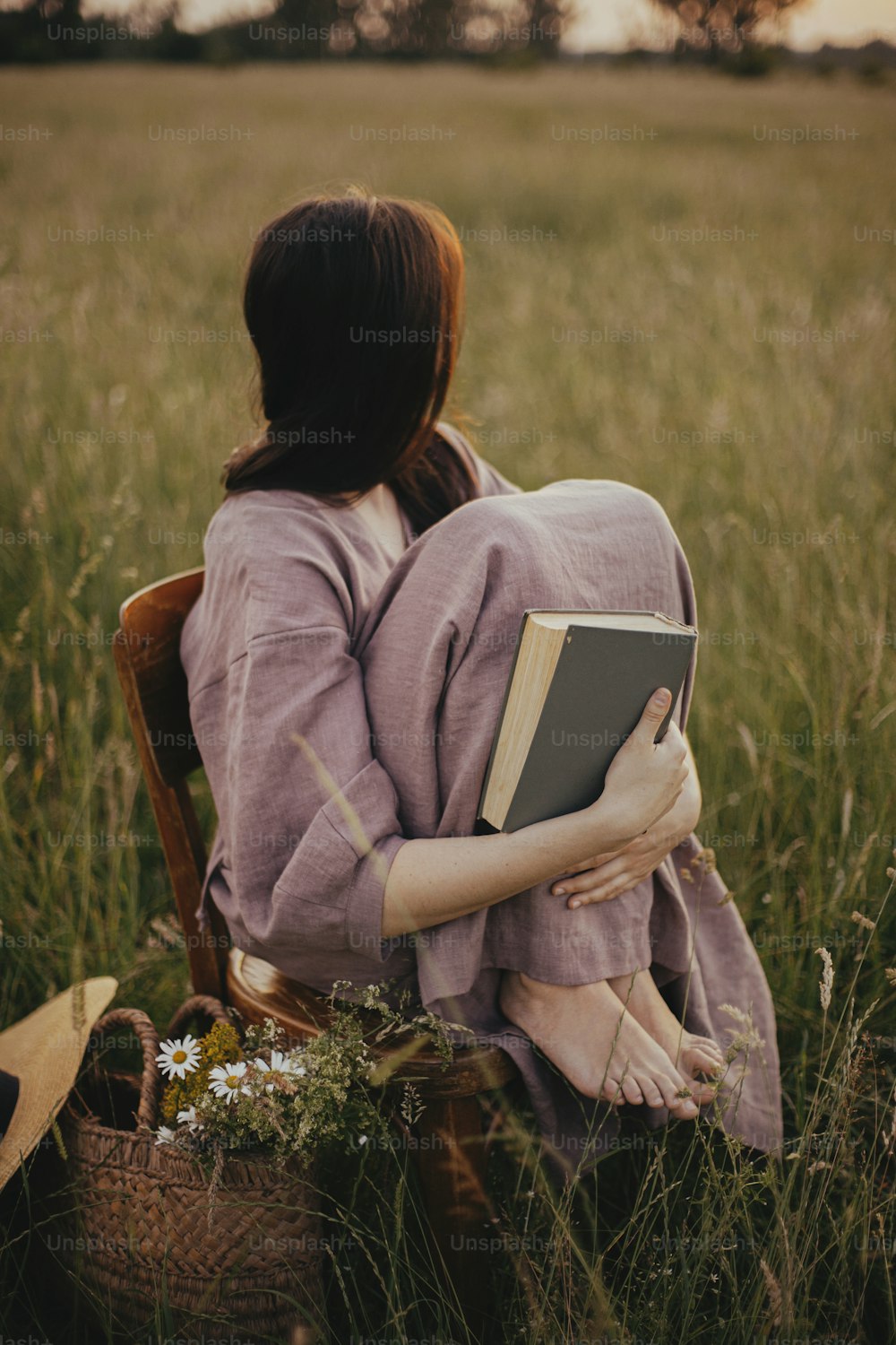 Beautiful woman in linen dress sitting on rustic chair and enjoying sunset in summer meadow. Young female relaxing with book and basket of flowers in countryside. Atmospheric tranquil moment