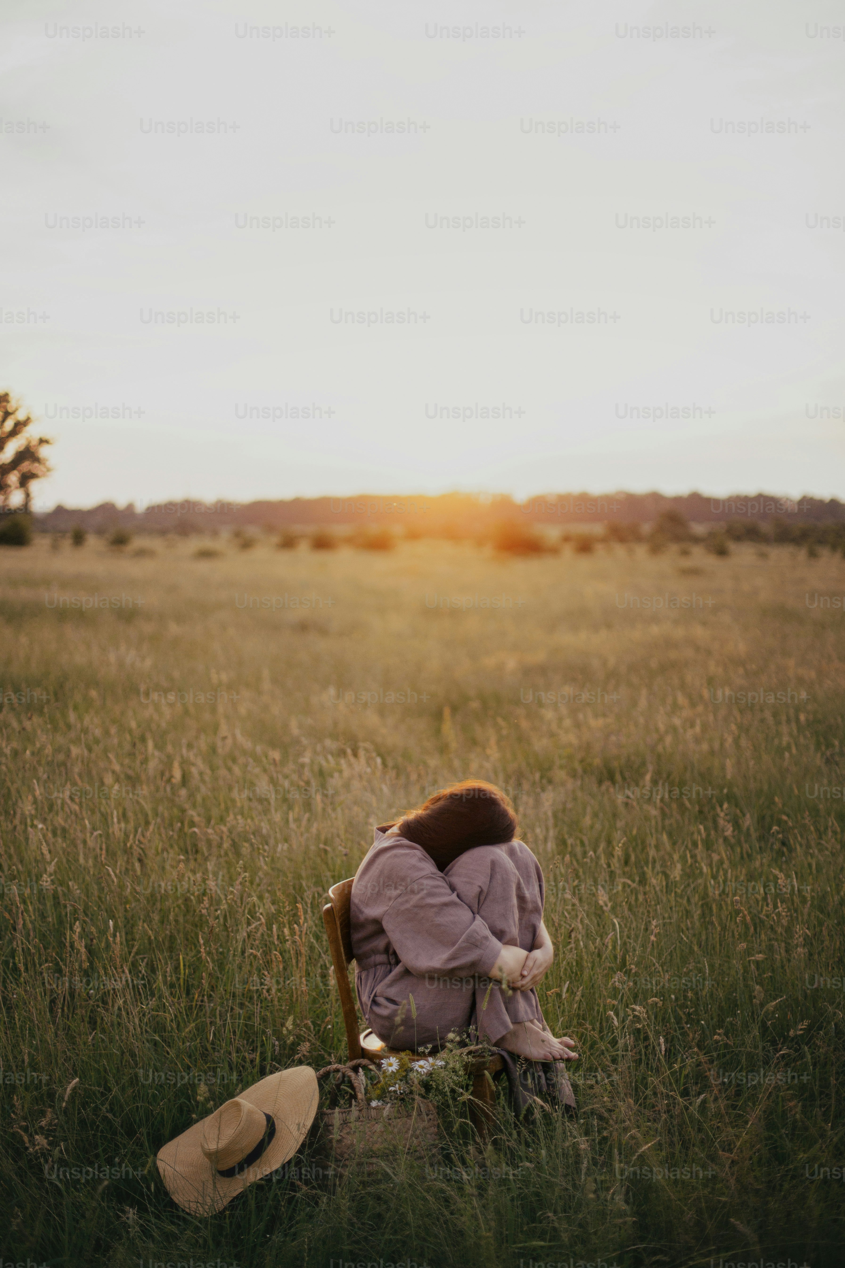 Beautiful woman in linen dress sitting on rustic chair and enjoying sunset in summer meadow. Young female relaxing with basket of flowers and hat in countryside. Atmospheric tranquil moment