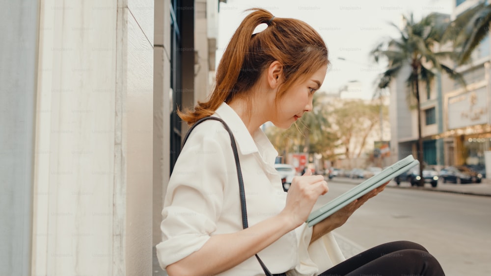 Successful young Asia businesswoman in fashion office clothes using digital tablet and typing text message while sitting alone outdoors in urban modern city in the morning. Business on the go concept.