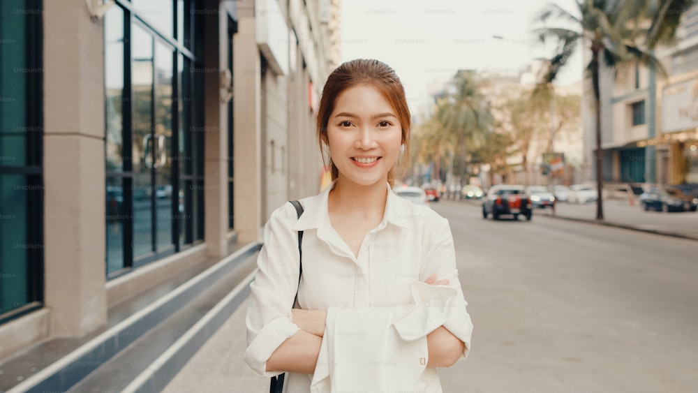 Erfolgreiche junge asiatische Geschäftsfrau in modischer Bürokleidung, die lächelt und in die Kamera schaut, während sie morgens glücklich ist, allein im Freien in der urbanen modernen Stadt zu stehen. Business on the go-Konzept.