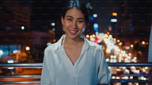 Successful young Asia businesswoman in fashion office clothes smiling and looking at camera while happy standing alone outdoors in urban modern city at night. Business on the go and commuter concept.