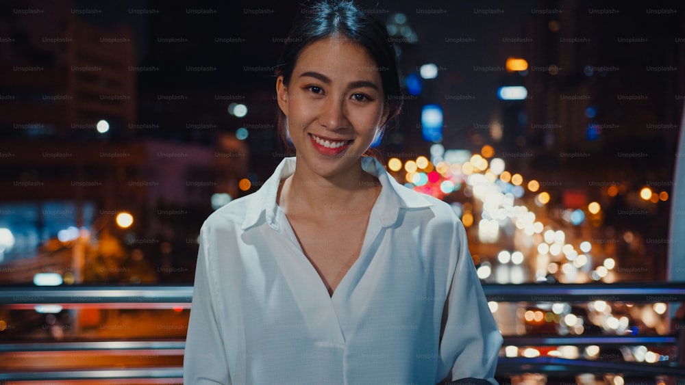 Erfolgreiche junge asiatische Geschäftsfrau in modischer Bürokleidung, die lächelt und in die Kamera schaut, während sie glücklich ist, nachts allein in der urbanen modernen Stadt zu stehen. Business on the go und Pendlerkonzept.