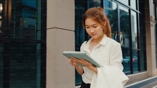 Successful young Asia businesswoman in fashion office clothes using digital tablet and typing text message while walking alone outdoors in urban modern city in the morning. Business on the go concept.