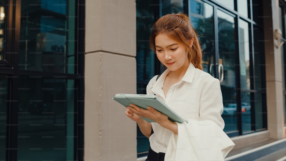 Successful young Asia businesswoman in fashion office clothes using digital tablet and typing text message while walking alone outdoors in urban modern city in the morning. Business on the go concept.