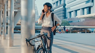 Asian businesswoman with backpack pick up mobilephone talk smiling watch smartwatch in city street go work at office. Sport girl use phone business. Commute to work by bike, Business commuter in city.