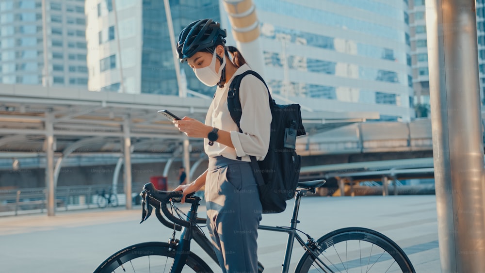 Asian businesswoman carry backpack wear anti virus protection mask take bicycle walk and check phone in city street go to work at office. commute to work, Business commuter for covid-19 concept.