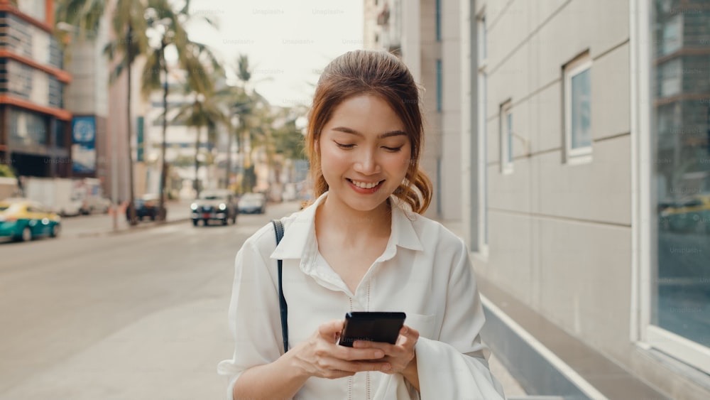 Successful young Asia businesswoman in fashion office clothes using smart phone and typing text message while walking alone outdoors in urban modern city in the morning. Business on the go concept.