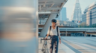 아시아 사업가는 사무실에서 일하러 가고 미소를 지으며 배낭을 메고 도시 거리의 건물 주변에서 자전거를 타고 돌아다닙니다. 자전거 통근, 자전거 통근, 비즈니스 통근 개념.