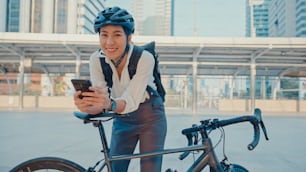 Sorride donna d'affari asiatica con lo zaino usa la fotocamera dello smartphone in città stand in strada con la bici vai al lavoro in ufficio. La ragazza sportiva usa il telefono per lavoro. Pendolarismo al lavoro, pendolare d'affari in città.