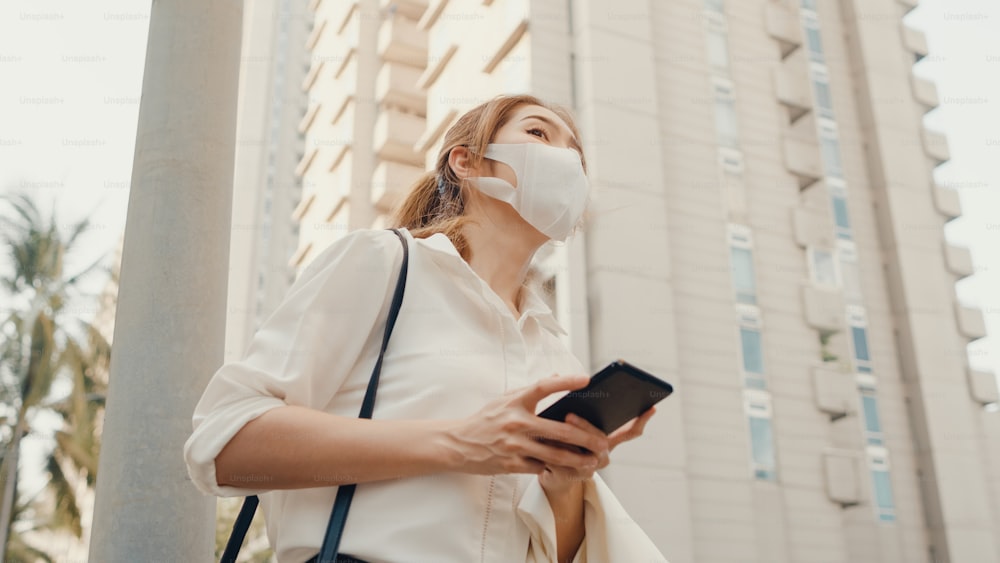 의료 얼굴 마스크를 쓴 패션 오피스 옷을 입은 젊은 아시아 사업가는 택시를 잡고 도시 현대 도시의 야외에 서 있는 동안 스마트폰을 사용합니다. 이동 중 비즈니스 개념.