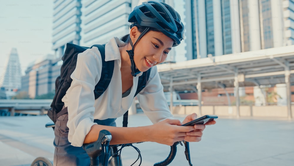 Sourire femme d’affaires asiatique avec sac à dos utiliser un téléphone intelligent regarder la caméra dans la ville se tenir dans la rue avec un vélo aller au travail au bureau. Une fille sportive utilise le téléphone pour le travail. Se rendre au travail, Trajet d’affaires en ville.