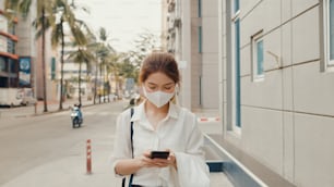 Erfolgreiche junge asiatische Geschäftsfrau in Mode-Bürokleidung trägt medizinische Gesichtsmaske mit Smartphone, während sie morgens alleine im Freien in der urbanen modernen Stadt spazieren geht. Business on the go-Konzept.