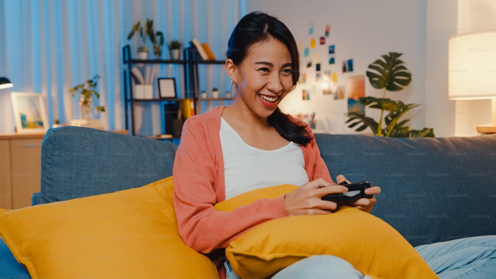Young Asia lady using wireless controller play video game having funny happy moment on sofa in living room at home night. Stay at house, Self quarantine activity for covid or coronavirus quarantine.