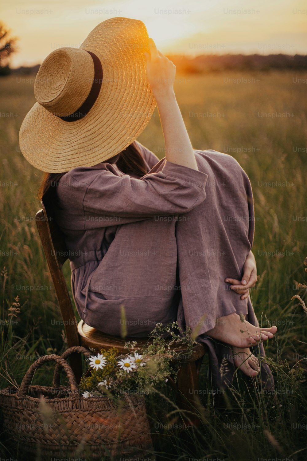 Mulher bonita no vestido de linho e chapéu de palha sentado na cadeira rústica com cesta de flores no prado de verão. Fêmea jovem apreciando o pôr do sol e relaxando no campo. Momento de tranquilidade atmosférica