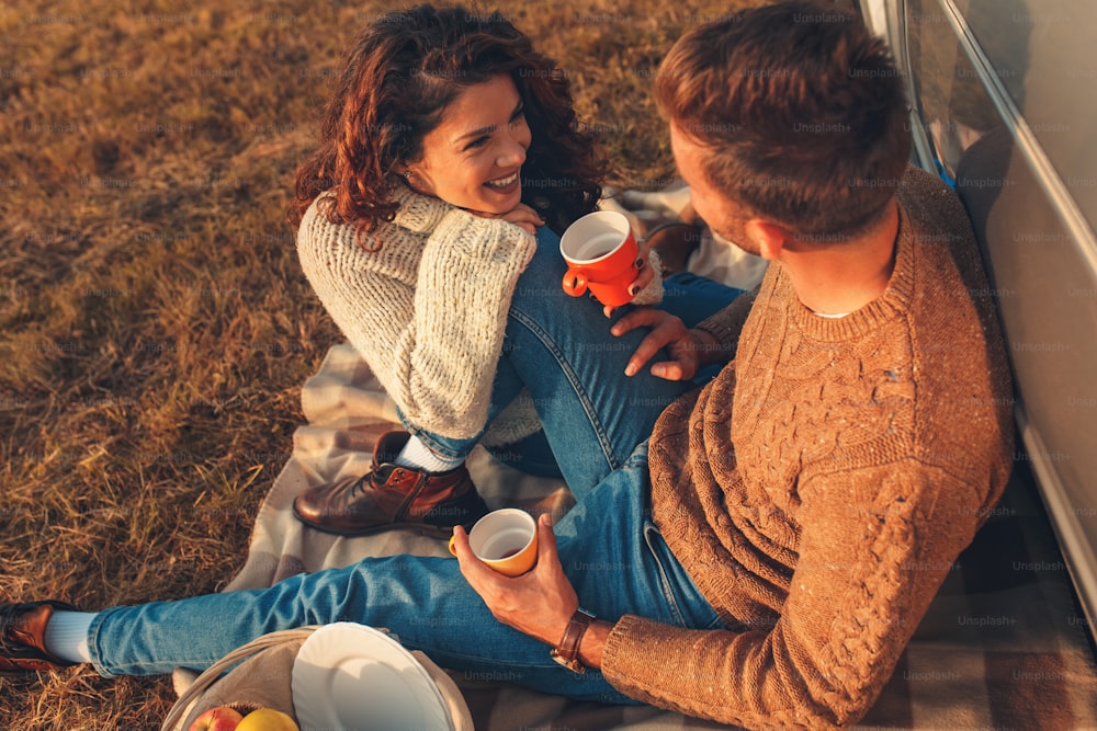 일몰에 피크닉 시간을 즐기는 아름다운 젊은 커플. 그들은 차를 마시고 구식 차에 기대어 초원에 앉아 있습니다.