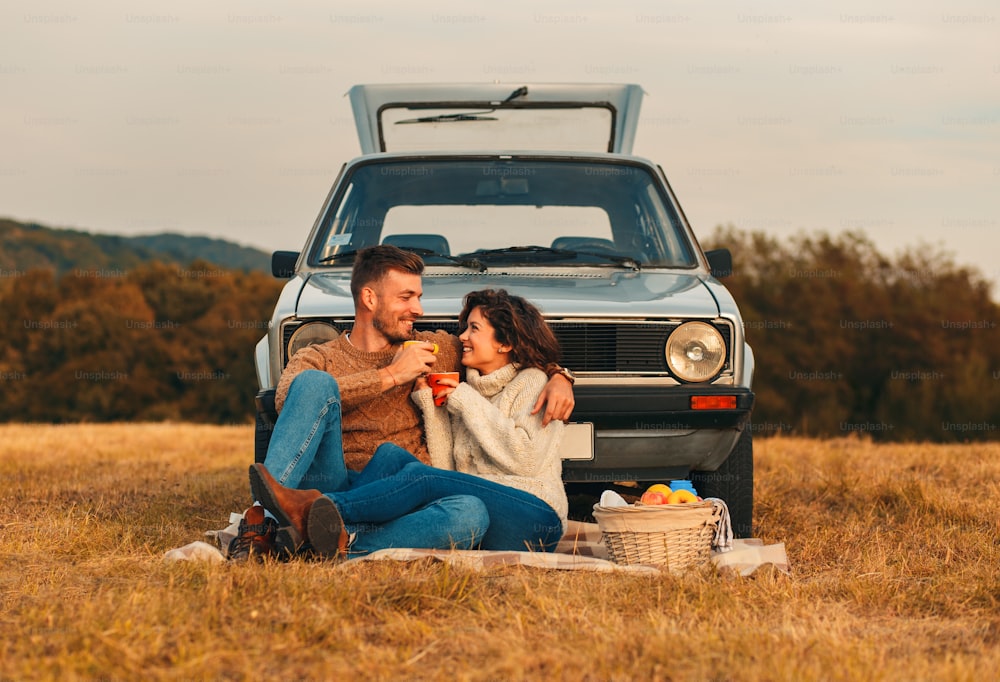 Beautiful young couple enjoying picnic time on the sunset. They drinking tea and sitting in a meadow leaning on old fashioned car.