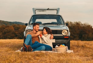 Beautiful young couple enjoying picnic time on the sunset. They drinking tea and sitting in a meadow leaning on old fashioned car.